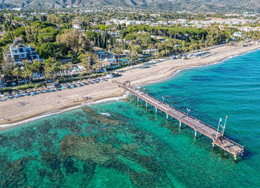 Aerial view of a picturesque coastal town with clear turquoise waters, a long wooden pier extending into the sea, and a beach lined with sun loungers and umbrellas. Nearby, there are lush green trees, residential buildings, and a backdrop of mountains.