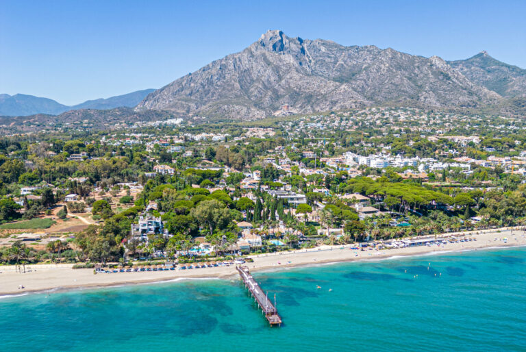 Aerial shot of a vibrant beachside community with crystal-clear waters, lush greenery, and a majestic mountain in the background, exemplifying high-quality media production in showcasing premium real estate locations.