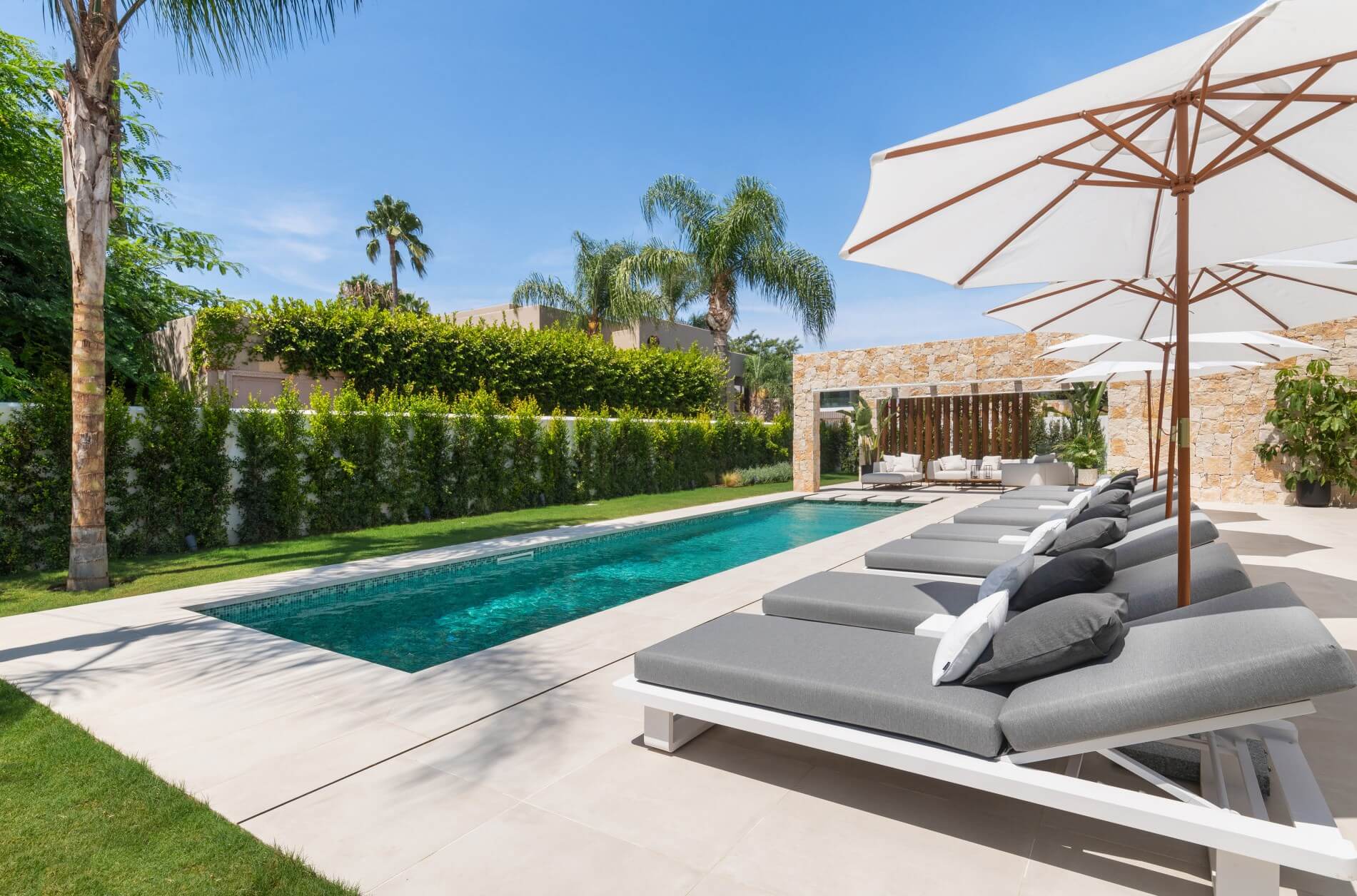 An upscale outdoor pool area with a row of stylish gray sun loungers shaded by large white umbrellas. The pool has a rectangular design and is surrounded by neatly trimmed hedges and a variety of lush palm trees. A stone wall feature with an integrated seating area is visible in the background, creating a luxurious and relaxing atmosphere.